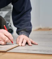 Hombre midiendo y marcando un panel de suelo de vinilo con un lápiz rojo, mientras trabaja en la instalación de un piso en una habitación.