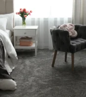 Habitación acogedora con cama blanca, almohadas suaves y una manta gris. Junto a la cama, mesita de noche con flores y una caja organizadora. Sillón gris elegante y tocador con productos de belleza. Suelo de alfombra gris claro.