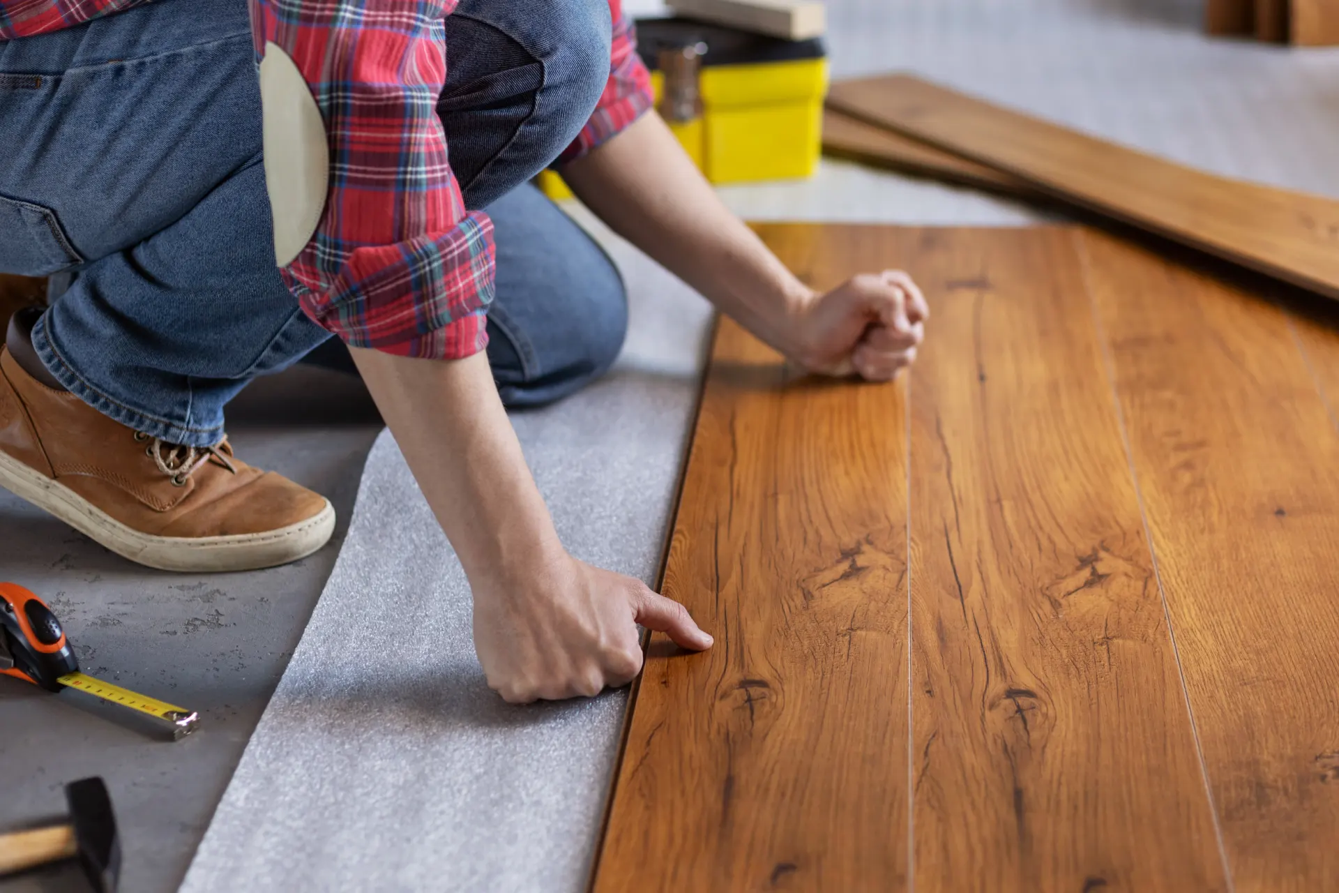 Persona instalando piso de madera en un espacio interior, utilizando herramientas como cinta métrica y martillo, mientras se asegura de que las tablas encajen correctamente.