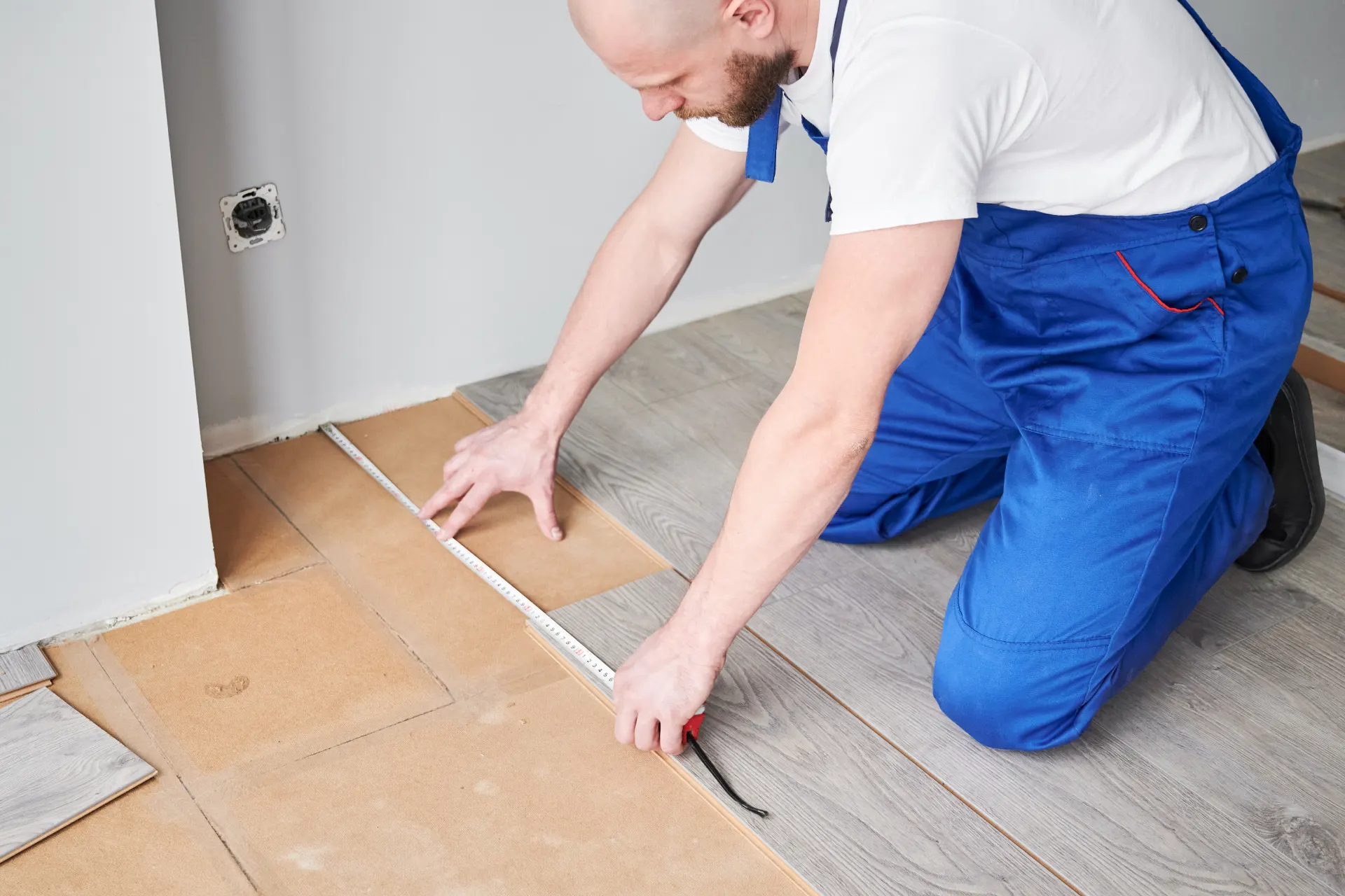 Trabajador midiendo el suelo para la instalación de pisos, usando una cinta métrica, en un ambiente interior con paneles de madera.